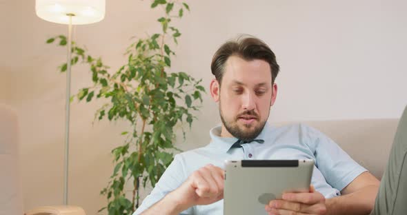 Young Bearded Man is Lying on the Sofa and Holding Tablet in Hands He is Relaxing and Scrolling the