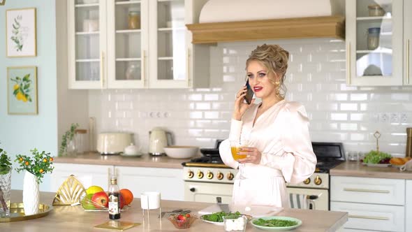 Beautiful Young Woman Talking on Phone in Kitchen Holds Glass of Fresh Juice in Her Hand