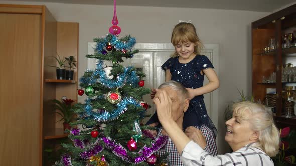 Kid Girl with Senior Grandmother and Grandfather Decorating Artificial Christmas Tree with Toys