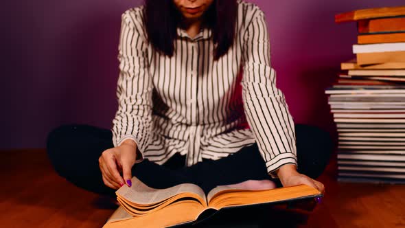 Young Woman Sitting on Floor with Pile of Book and Flipping Through Book on Purple Background