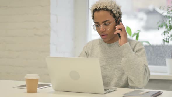 Angry African Woman Talking on Smartphone While Using Laptop in Office