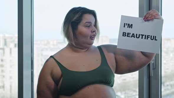 An Overweight Woman Holding a Nameplate with a Sign I'M BEAUTIFUL and Eating a Burger