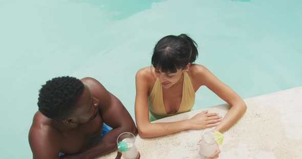 Happy diverse couple wearing swimming suits and drinking drinks at swimming pool in garden