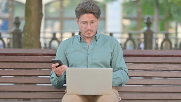 Middle Aged Man Browsing on Smartphone while working on Laptop