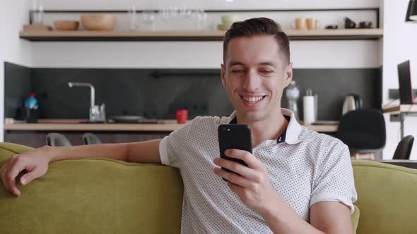 Close Up of Cheerful Young Man Sitting on the Sofa Using Smartphone Smile in the Modern Apartment