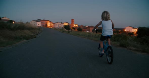 Child Boy Ride on Bicycle on Empty Country Road with Village Light on Background