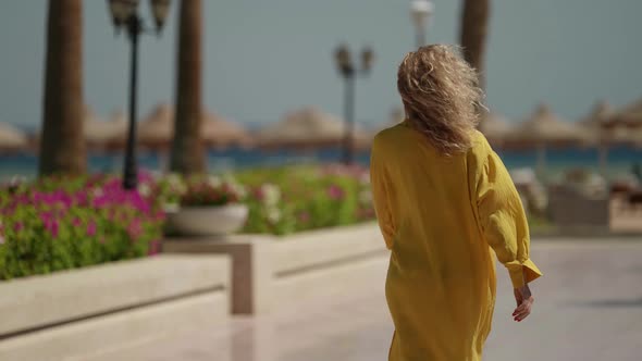 Blonde Woman in Yellow Dress is Walking to Sea Over Territory of Luxury Hotel