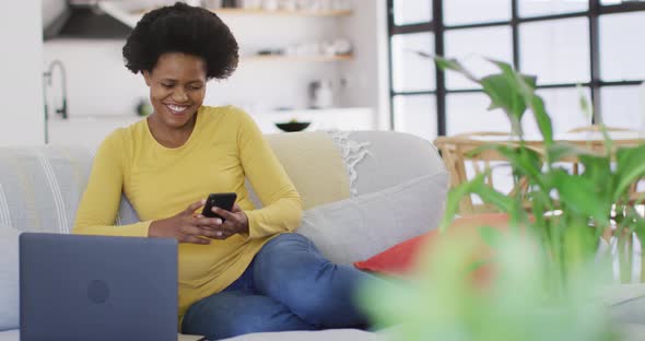 Happy african american woman sitting on sofa using laptop and smartphone