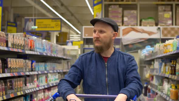 A Bearded Man in a Cap with a Shopping Cart Walks Through the Rows of Various Goods Chooses a