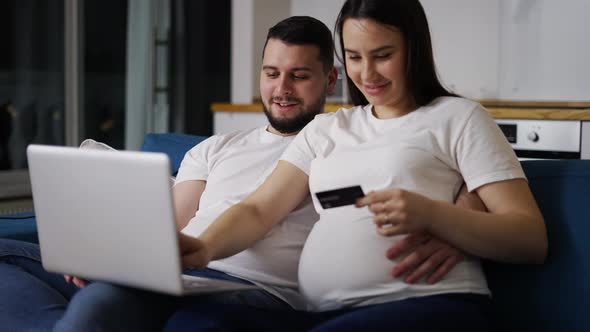 Couple Expecting Baby Sitting on Sofa at Home and Shopping Online Using Laptop