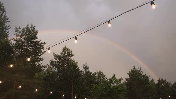 Bright Rainbow Over Green Forest in Overcast Sky with Garland of Glass Bulbs