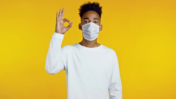 Mixed Race Man in Protective Medical Mask Showing Thumb Up Sign Over Yellow Background. Positive