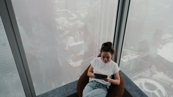 An Attractive Girl Sits on a Soft Pouf By the Window Against the Backdrop of a City of Skyscrapers
