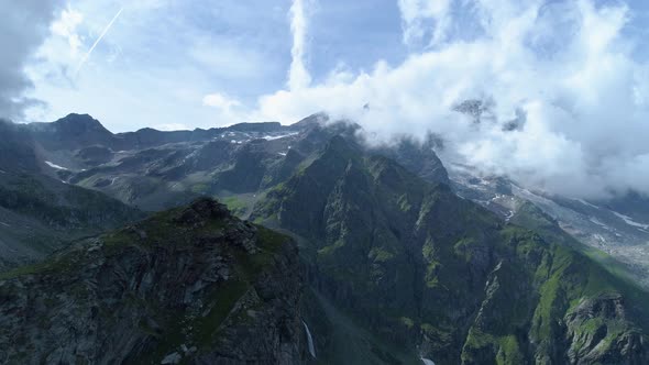 Moving Forward Over Majestic Alpine Mountain Peak Revealing Valley in Sunny Cloudy Summer