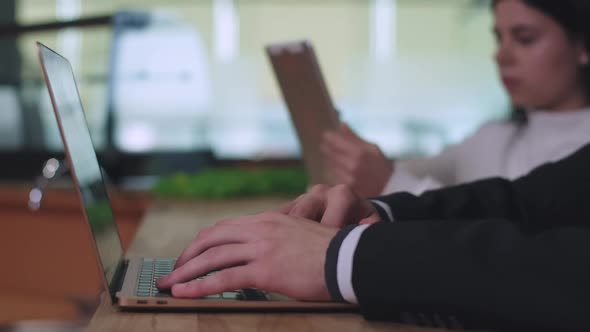 Male Caucasian Hands Typing on Laptop Keyboard with Blurred Woman Using Tablet at the Background