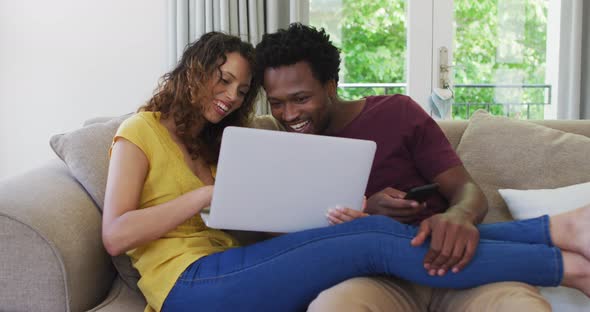 Happy biracial couple sitting on sofa with laptop and laughing