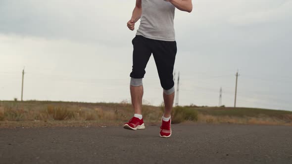 Man in Sportswear and Sneakers Confidently Runs Along Asphalt Road Against Background of Field Front