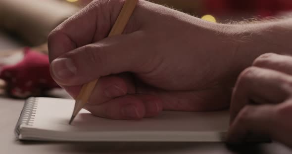 Closeup Man Writes Christmas Gifts in Notepad with a Pencil Under Warm Light in the Evening