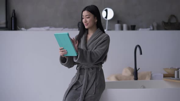 Portrait of Slim Charming Asian Woman in Bathrobe Sitting on Bathtub Surfing Social Media in Slow