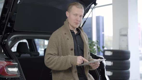 Young Caucasian Man Standing at Car Trunk Using Tablet, Looking at Camera and Smiling, Confident