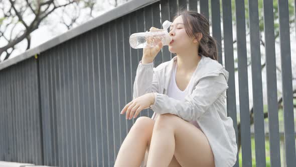 4K Asian woman drinking water from a bottle while jogging at public park in the morning.