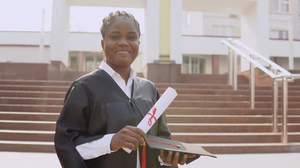 Young African American Female Graduate Standing in Front of the Camera with a Diploma and a Hat in