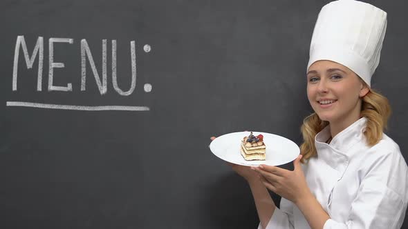 Friendly Lady Cook Holding Cake on Plate, Menu Written in Blackboard, Horeca