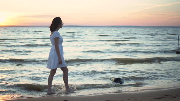 Attractive Young Woman Walks Along the Sea Beach at Sunset. A Girl in a White Dress Walks Barefoot