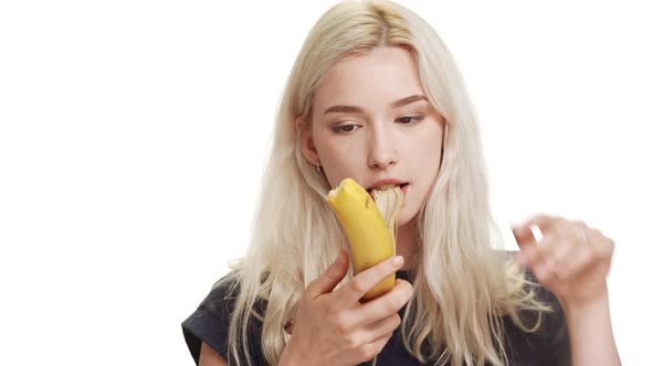 Young Caucasian Blonde Teenage Girl Cleaning and Eating Banana Covering Mouth with Hand on White