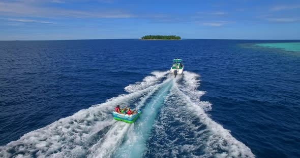 Aerial drone view of man and woman on an inflatable tube towing behind a boat to a tropical island