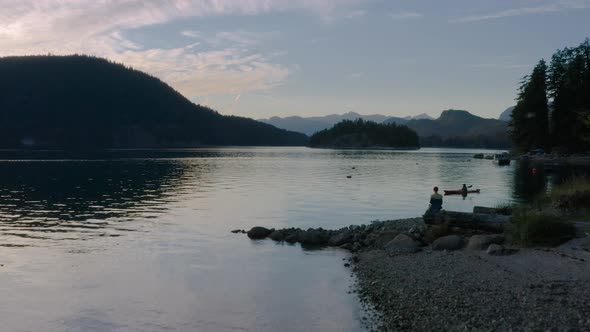 Drone Flying Towards A Woman Sitting On The Log At The Shore With Person Boating In Sechelt Inlet Ne