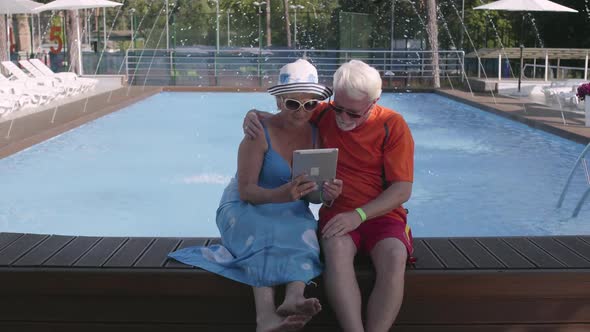 Happy Mature Couple Sitting on the Edge of the Pool with Tablet in Hands