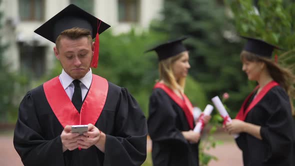 Happy Male Graduate in Mantle Choosing Hotel for Summer Holidays on Smartphone