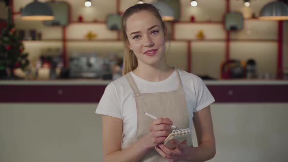 Portrait of Positive Caucasian Waitress Looking at Camera and Smiling. Young Woman in Apron Standing