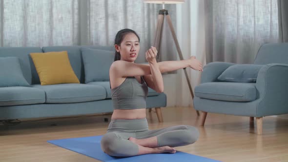 Asian Athletic Female In Sports Clothes Sitting On A Yoga Mat And Doing Shoulder Cross-Arm Stretch