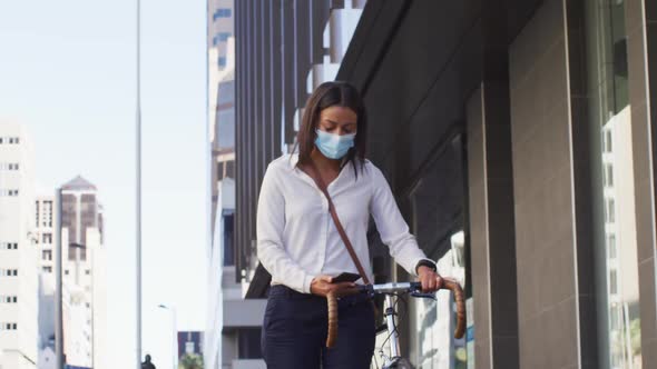 African american woman wearing face mask using smartphone wheeling bicycle