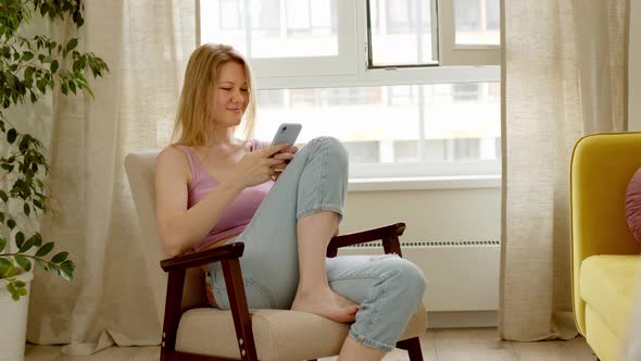 Beautiful Blonde Sits on Gray Retro Armchair in Pink Top and Jeans and Use Phone Typing Text