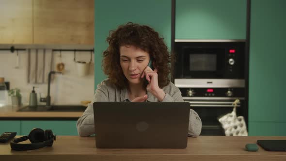 Young Curly Caucasian Woman is Talking on Her Phone in Front of a Laptop Computer Indoors