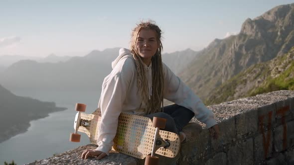 Portrait of happy dreadlocks woman holding longboard standing on mountain