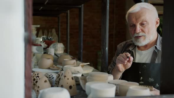 Elderly Potter in Dirty Apron Counting Pots in Workshop Writing Information in Notebook