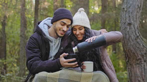 Young Happy Couple of Tourists in Love Sit Hugging in Nature Drinking Hot Tea From Thermos to