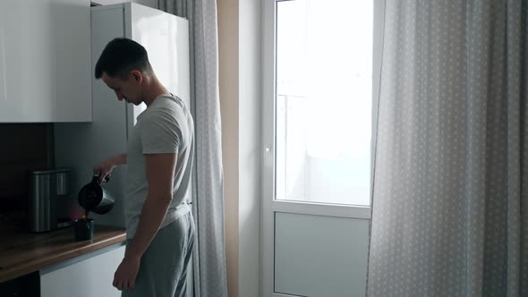 Handsome Young Man Pours Coffee From Coffee Maker and Walks Up to Window in Kitchen