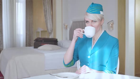 Portrait of Charming Caucasian Stewardess in Blue Uniform Drinking Coffee in the Morning in Hotel