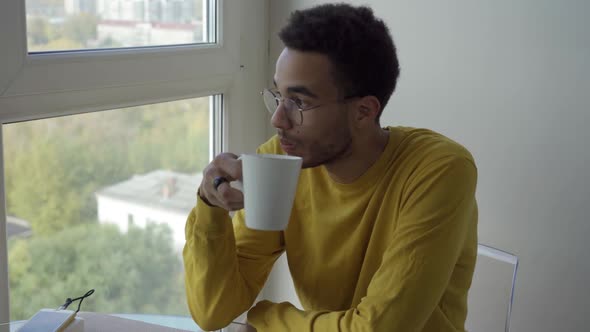 A Young African Man is Drinking Coffee While Sitting By the Window