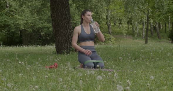 Woman Dressed in Leggings and Top Drinking Water in the Background of the Park