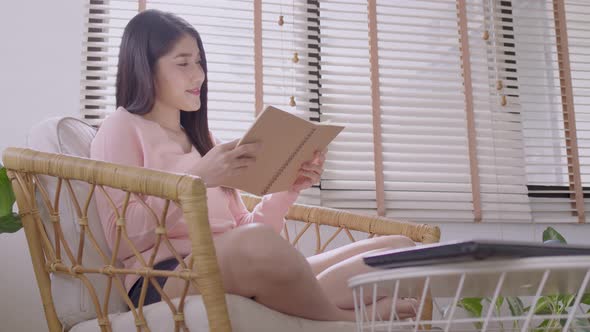 Asian woman reading at home sitting on modern chair relaxing in her living room reading book.