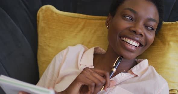 Happy african american woman lying on sofa in living room, writing in notebook