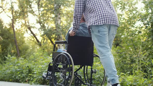 Man Pushes Wheelchair with His Beloved Disabled Woman on Walk in Park Back View Low View