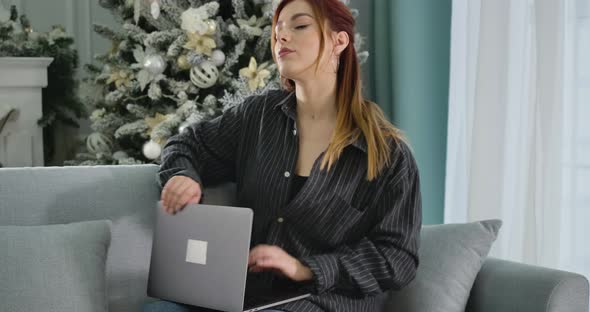 Charming Redhead Woman Closing Laptop and Stretching with New Year Tree at the Background. Portrait