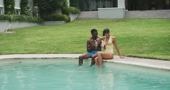 Happy diverse couple wearing swimming suits and drinking drinks at swimming pool in garden